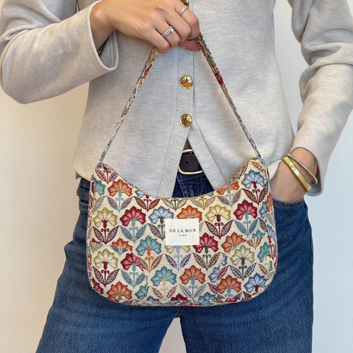 Model holding the DE LA MUR Flora floral shoulder bag over jeans, lifestyle shot
