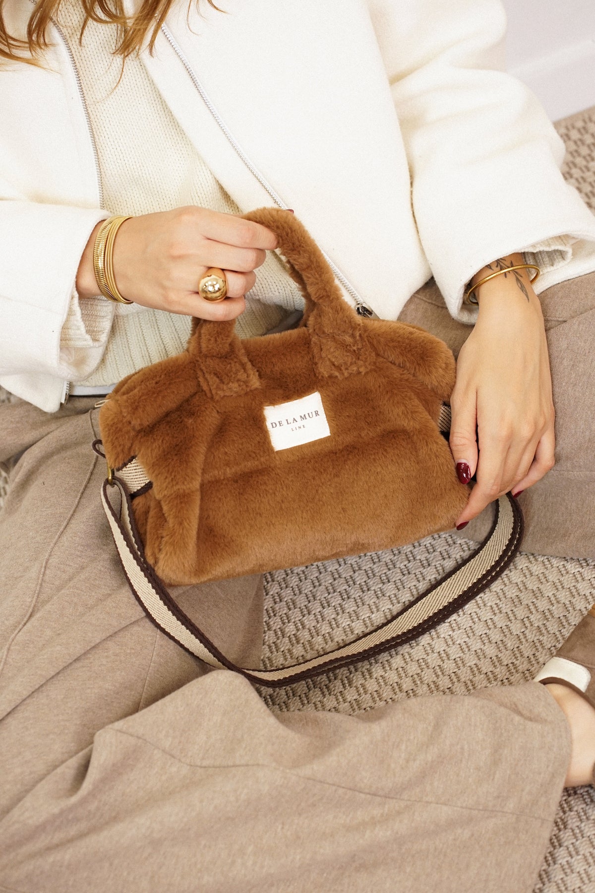 DE LA MUR camel brown faux fur small handbag with striped strap, held by a seated model