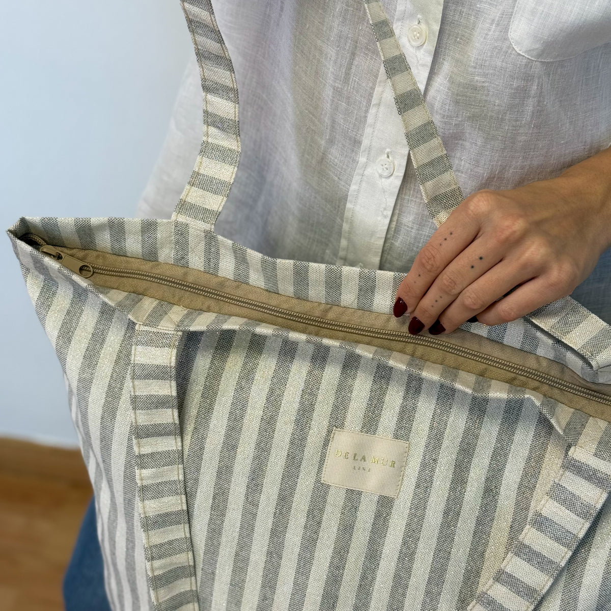 Model opening the DE LA MUR gray striped tote bag, showing zip detail