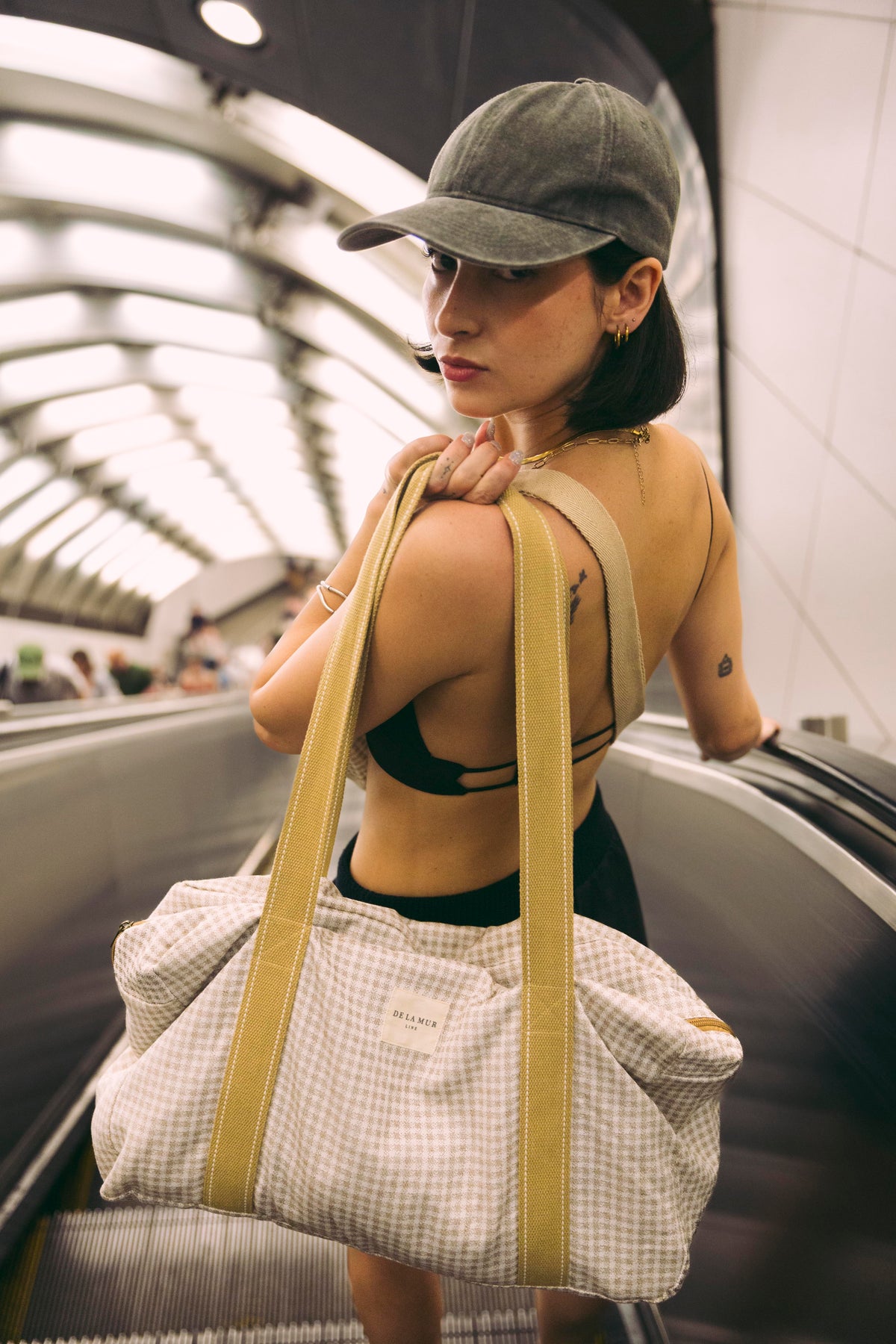Model on an escalator carrying the DE LA MUR Vison gingham jacquard sports bag