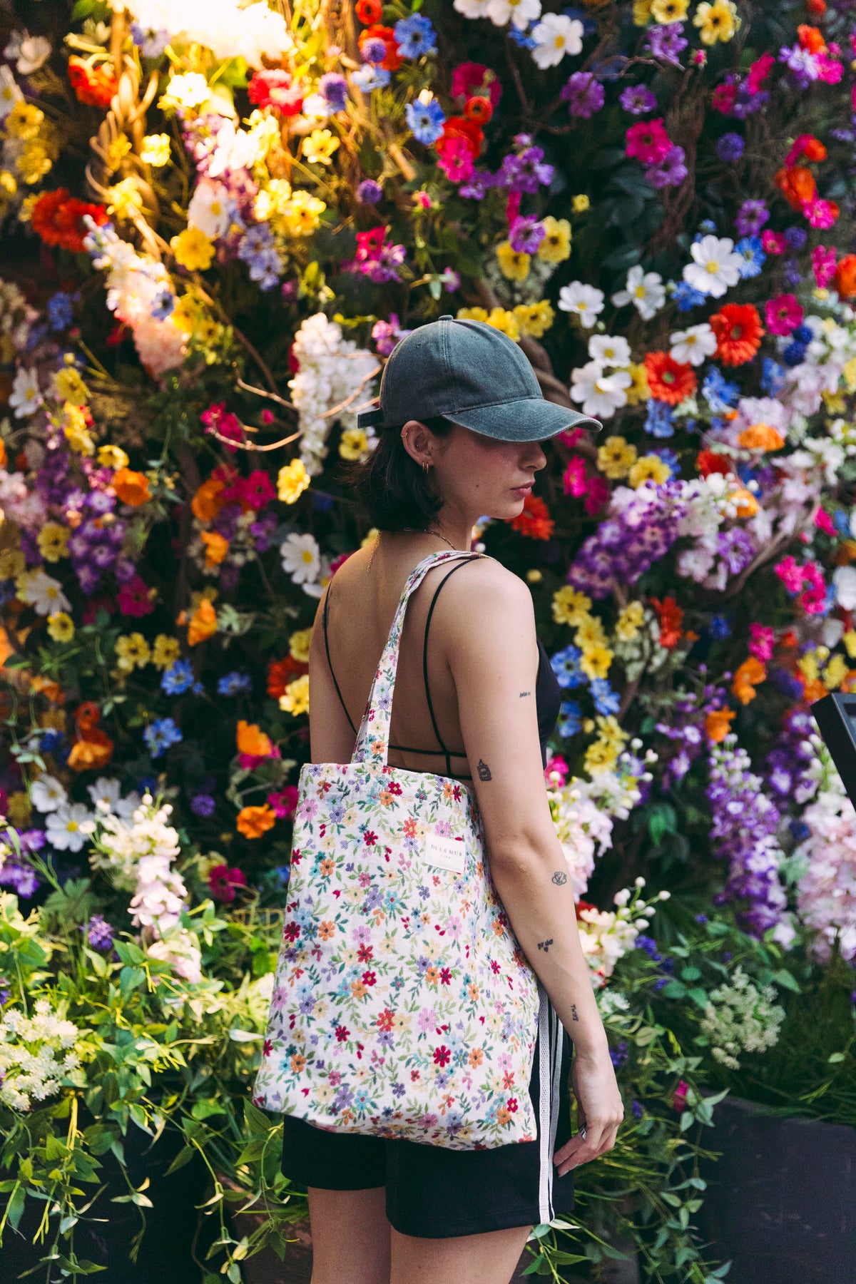 Model carrying the DE LA MUR Blum floral medium zippered tote bag on the shoulder, side view, by a flower wall