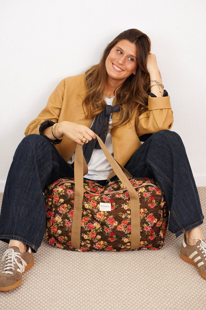 Seated model holding DE LA MUR Pepi floral jacquard sports bag in brown and red, lifestyle shot